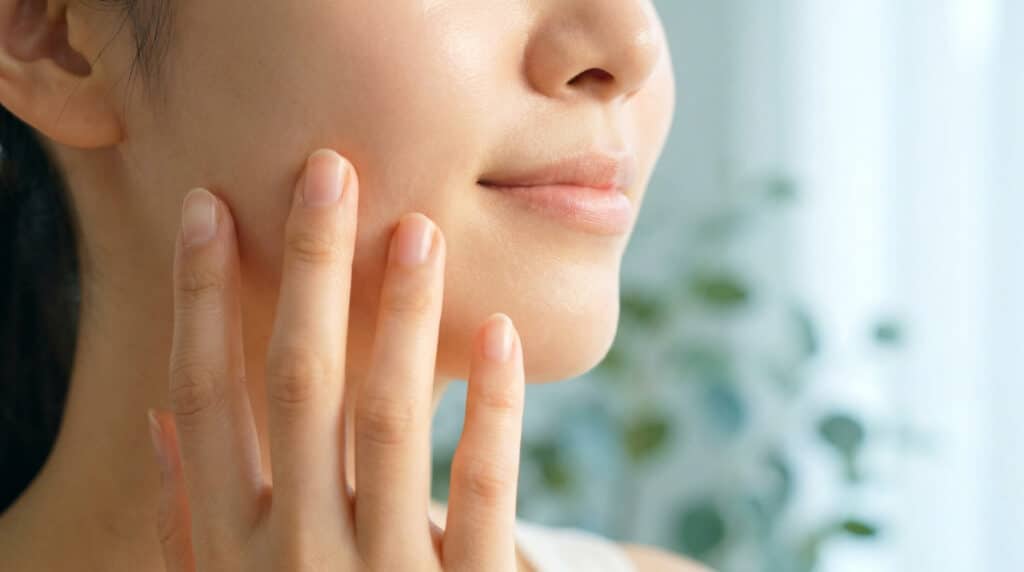 Gros plan sur le bas du visage d'une jeune femme touchant délicatement sa joue, peau nette et lumineuse.